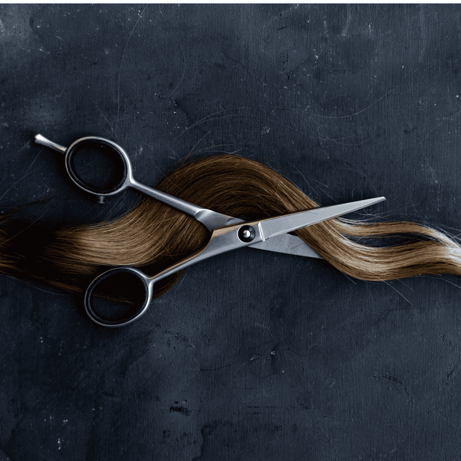Scissors cutting a strand of brown hair on a dark surface.