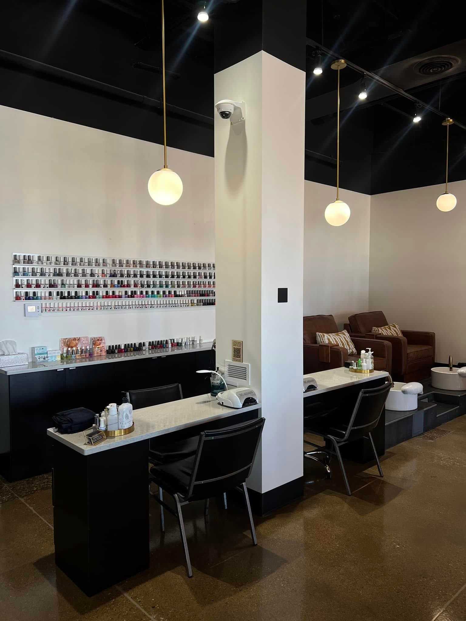 Modern nail salon with manicure stations, wall of nail polish, and pedicure chairs in the background.