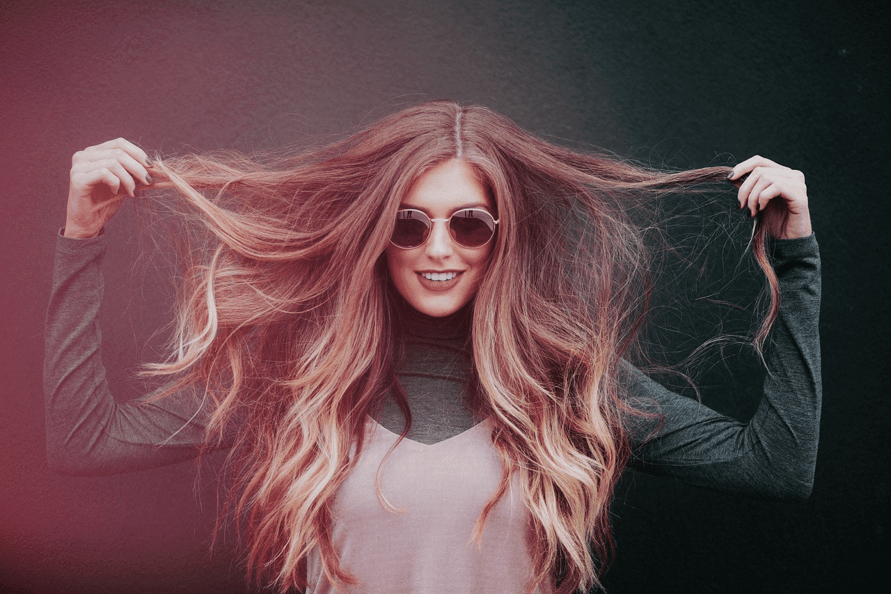 Young woman in sunglasses holding her long hair, smiling against a dark background.